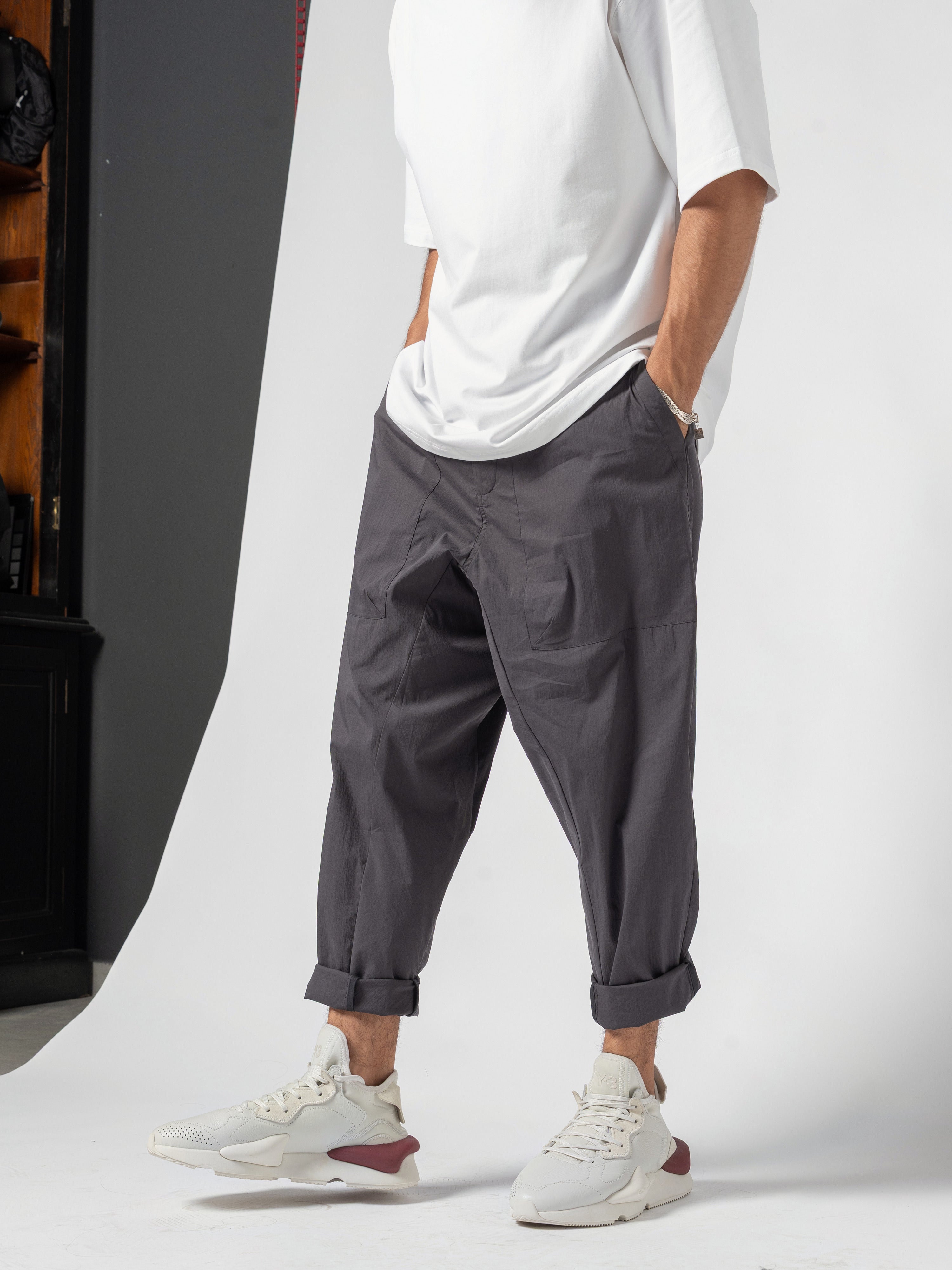 AITO Pleated Drop-Crotch Pants in Dark Grey - front three-quarter worn with a white tee, one hand in the side pocket, cuffs turned up over white trainers
